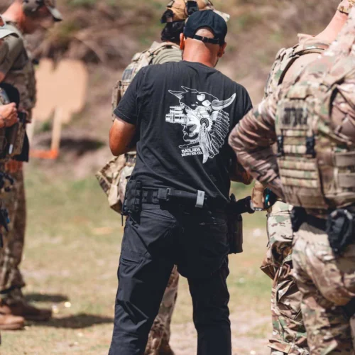 Back view of a man in a black graphic-t-shirt and cap, standing among soldiers in camouflage during outdoor training. He wears a duty belt with gear.