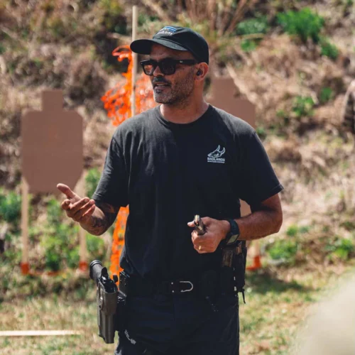 Man in a black shirt and cap instructs at an outdoor shooting range, gesturing while holding a handgun with holster nearby.