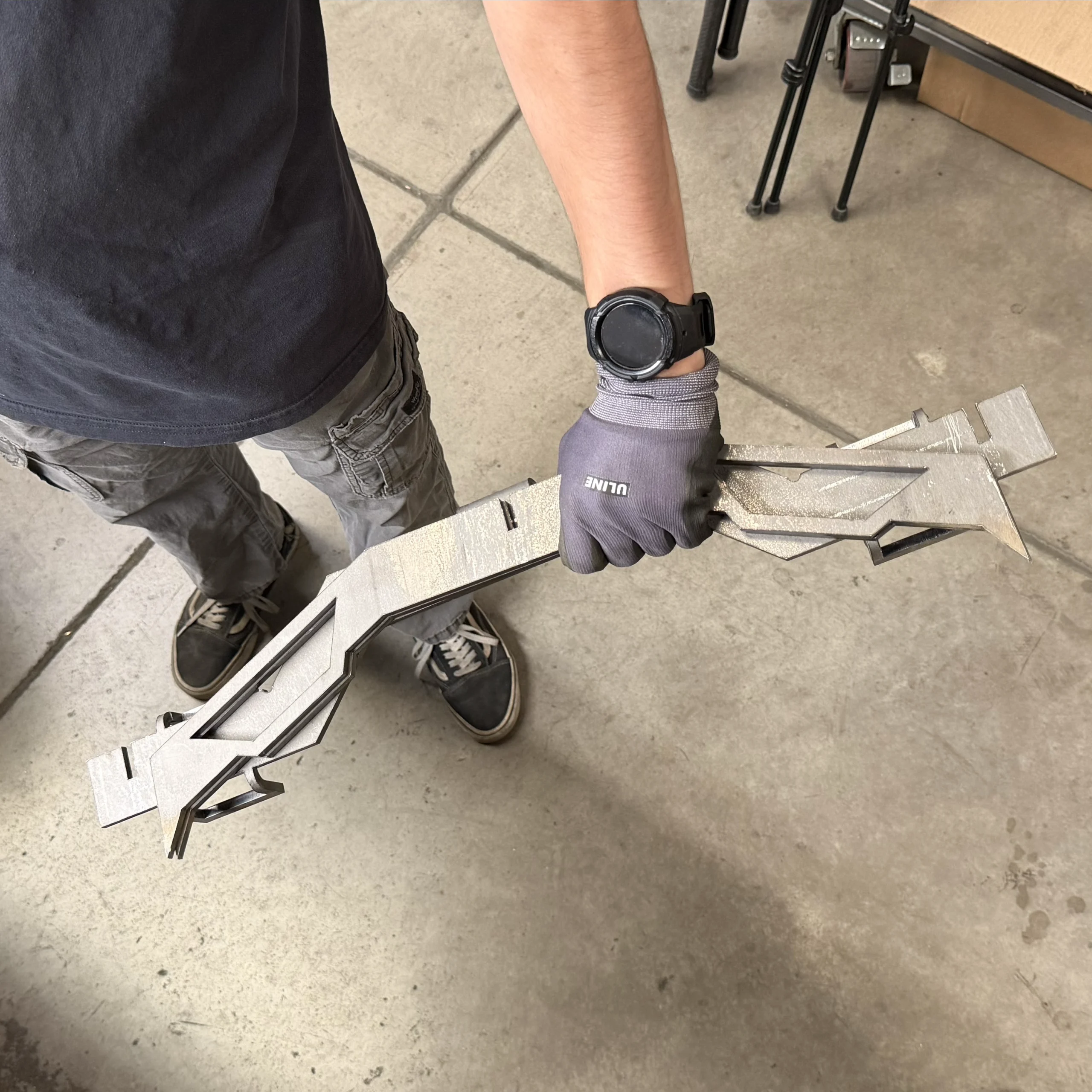 STAND – Badlands Munitions Co Person in a dark shirt and gloves holding a long, textured metal framework on a concrete workshop floor.