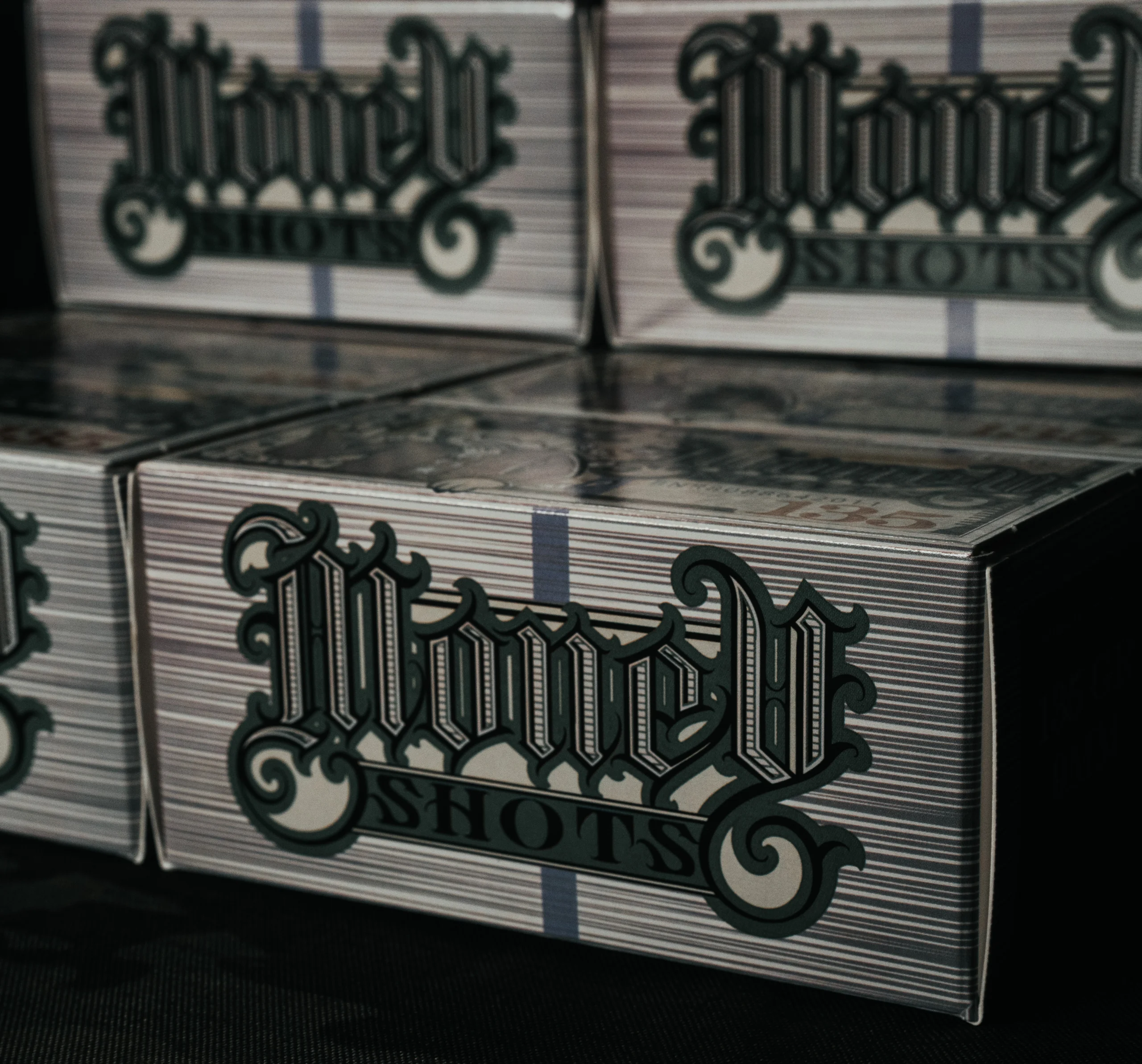 MONEY SHOTS BOX 2 – Badlands Munitions Co Close-up of stacked white boxes with dark teal ornate script lettering on a dark surface, conveying vintage packaging design.