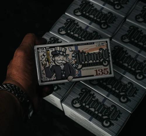 Close-up of a hand holding a decorative $135 bill featuring Uncle Sam, with stacks of similar bills in the dark background.