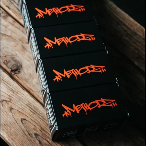 Close-up of black packaging boxes stacked on a wooden surface, featuring bright orange graffiti-style lettering.